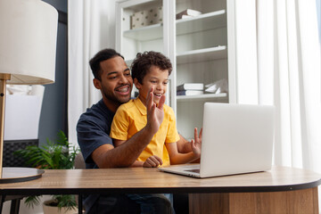 African American man and his son use laptop at home at the table, father and child communicate via video call online and greet