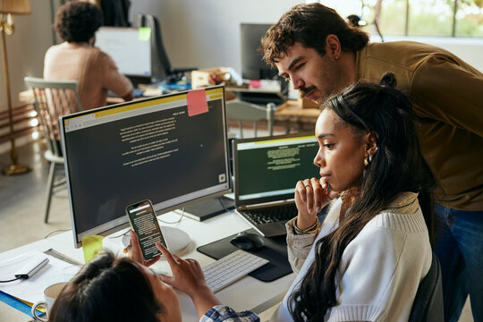 Male and female tech computer programmers looking at smart phone shown by colleague in office