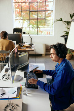 High angle view of young male programmer wearing wireless headphones working on computer while sitting at desk in office