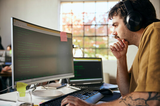 Focused male programmer wearing wireless headphones coding on computer in startup office