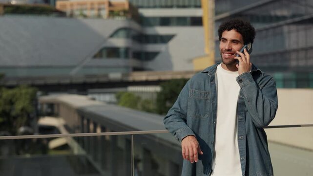 Portrait of young Hispanic man talks on smartphone and leans onto glass railing on street slow motion. Latin American guy calls via mobile phone as stands on footbridge in city