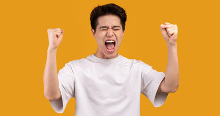 Winner Concept. Studio portrait of overjoyed asian man yelling with clenched fists raised up in the air, isolated over orange background. Excited guy cheering and screaming, celebrating victory