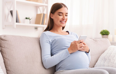 Obraz premium Happy Pregnant Woman Texting On Cellphone Sitting On Sofa At Home. Pregnancy Apps Concept