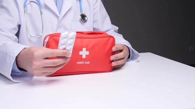 Medical professional wearing a white lab coat and stethoscope holding a blister pack of pills, with a red first aid kit resting on a white desk, representing healthcare and emergency treatment
