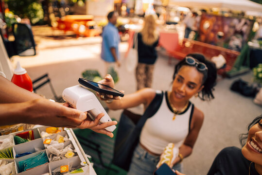 Hand of salesman holding credit card reader while female customer tapping smart phone on it at food truck