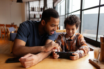 African American man and his son use smartphone in cafe at table, teenager and his father sit in restaurant and play video games on mobile phone