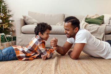 African American man competes with his son in arm wrestling at home on the carpet, teenager tests his strength with his father and smiles