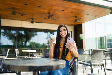 Happy woman smiling, holding coffee cup, looking at her smartphone at an outdoor cafe. Enjoying a social media break