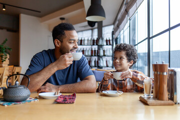 African American man and his son are having lunch in cafe at table, teenager and his father are sitting in restaurant and eating and drinking coffee