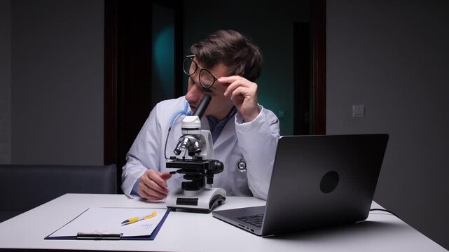 Doctor in white lab coat and stethoscope examining a sample with a microscope while sitting at a desk with an open laptop, conducting scientific research in a laboratory environment at night