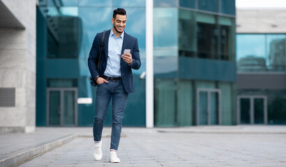 Confident arabic guy programmer with backpack walking by street, checking his mobile phone, chatting with friends, going home from office, panorama with copy space, full length