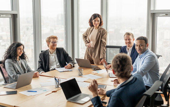 A group of diverse business professionals are seated around a large conference table in a modern office space. One woman stands at the head of the table, while the others focus on the discussion