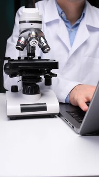 Vertical video scientist in lab coat analyzing samples with a microscope and simultaneously typing on a laptop, combining traditional research methods with digital data analysis