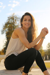 Happy beauty face woman with long hair looking in camera sitting in casual clothes on stairs outdoor summer bright sun background. Closeup fun relaxing person street portrait. Natural light