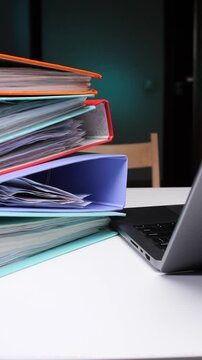 Vertical video stacked office binders and scattered documents beside a laptop on a white desk, conveying busy administrative work and organization