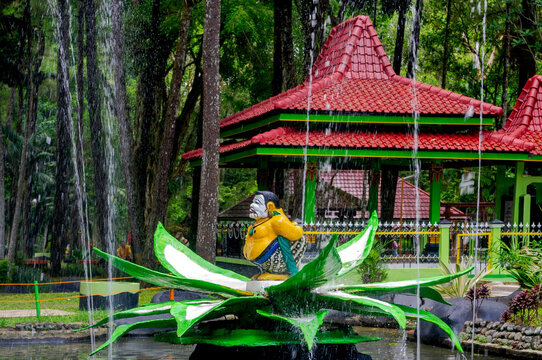 A Semar wayang statue sits in a garden fountain, in a public park, during the day, representing Javanese wisdom, as water flows gently around the sculpture.