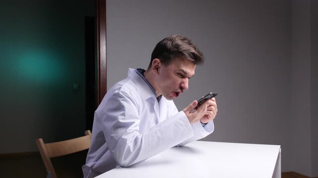 Doctor in medical gown and lab coat sitting at desk, angrily reacting to bad news on a smartphone, shouting in frustration and stress over a workplace crisis and communication failure