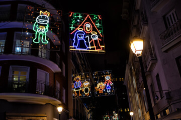 Colorful Christmas light displays hanging above a city street at night, showing festive figures and nativity scenes, creating a joyful holiday atmosphere in an urban setting