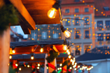 Festive scene with numerous glowing Edison-style string lights draped over wooden structures on European Christmas market. Backdrop of historic buildings with lights adds to cozy holiday atmosphere