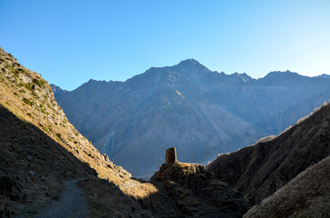 Deep mountain gorge under clear blue sky. Small, ancient stone watchtower stands in the center foreground against towering hazy mountain peak in the background. Kazbegi, Caucasus, Georgia