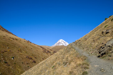 Mountain trail ascends between two dry, grassy slopes under a deep blue sky, perfectly framing the distant, snow-capped peak of Mount Kazbek in the center background. Georgia