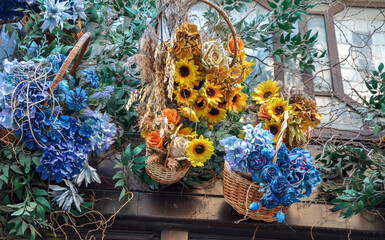 Wicker baskets filled with vibrant flowers including clusters of blue hydrangeas, yellow sunflowers, and roses&mdash;hang on a wooden structure, surrounded by green foliage against a window backdrop

