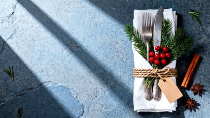 Rustic holiday table setting with napkin-wrapped cutlery, evergreen sprigs, and red berries on a blue stone surface