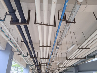 This photo shows a bottom-up view of a building's ceiling, lined with pipes and utility lines. Rows of white, black, and blue pipes and rusted metal pipes hang from iron supports, accompanied by fluor