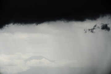 Dramatic dark storm clouds gather over a distant mountain range and lush landscape, with a bright horizon line below. Moody, extreme weather.