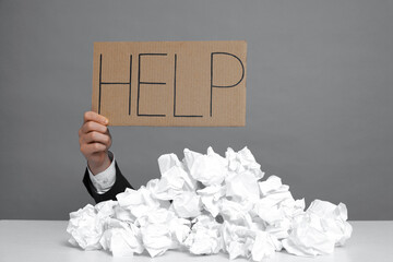 Businessman holding sign with word Help at white table with pile of crumpled paper balls against...