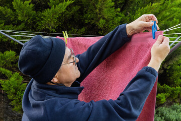 Senior man wearing a hat hangs a pink towel on an outdoor rotary washing line using pegs. Domestic laundry, everyday life, sustainability, and outdoor household chores