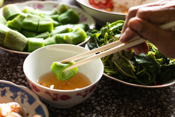 Close up of a hand using bamboo chopsticks to dip a fresh boiled luffa slice into fish sauce, typical of rustic Vietnamese cuisine