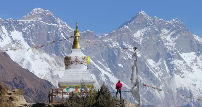Slow telephoto orbit drone shot of a male tourist standing at the Namche memorial viewpoint, taking photos with his phone. Majestic Himalayan backdrop along the Everest Base Camp trek, Nepal.