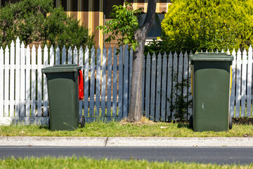 Residential wheelie bins placed on a suburban street verge in front of a white picket fence and house in Australia. Concept of household waste management, local council services, curbside collection