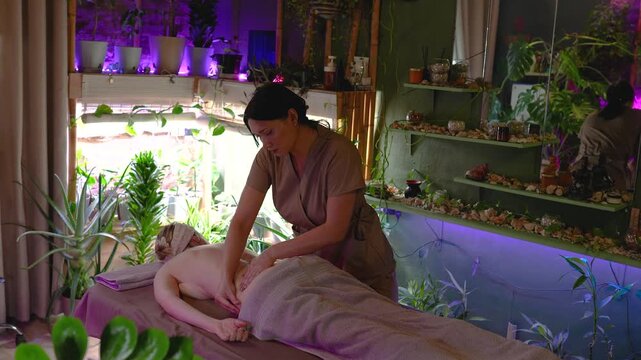 Beautiful half naked woman lying on bed under towel in authentic cosmetology spa salon with relaxing atmosphere for full body treatment, lady receiving lower back massage from professional therapist.