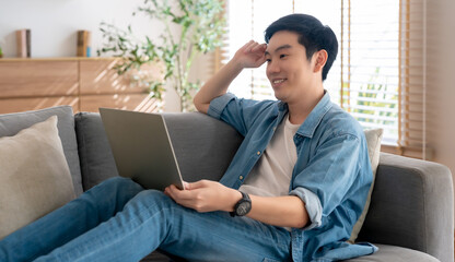 Young Asian man relaxing on comfortable couch, using laptop at home, happy boy chatting with friends in social network, spending weekend, watching movie, shopping online