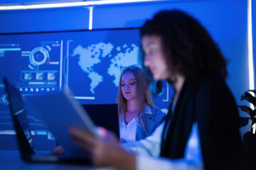 Women working together in a modern office setting with digital screens and blue lighting at night