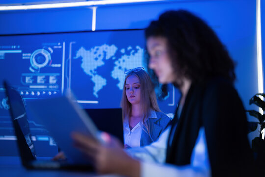 Women working together in a modern office setting with digital screens and blue lighting at night