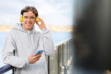 Fototapeta premium Young man in grey hoodie with headphones and mobile phone on embankment