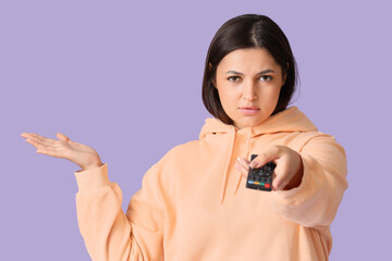 Upset young woman with remote controller watching TV on lilac background