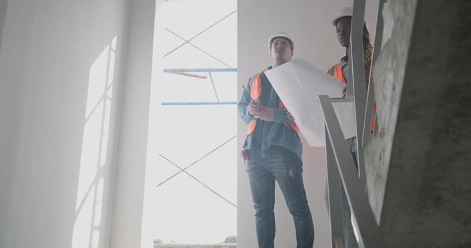 Engineer discuss blueprint at construction site, making decisions for architectural plan on upper floor. Both are dressed in safety gear. Collaborative approach to ensure project accuracy and progress