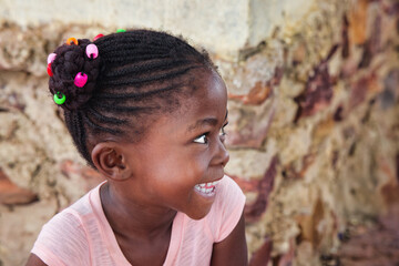 village happy african girl child profile portrait, cornrows braids dreadlocks hairstyle, big toothy smile, outdoors in the nature ,stone wall behind