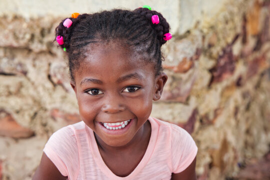 village happy african girl child portrait, cornrows braids dreadlocks hairstyle, big toothy smile, outdoors in the nature ,stone wall behind