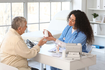 Young African-American doctor giving pill bottle to senior woman at table in clinic