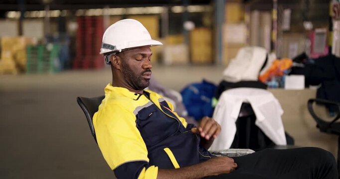 African American male warehouse staff resting with eyes closed on break wearing hardhat and uniform while seated near inventory and packaging area in large industrial storage facility