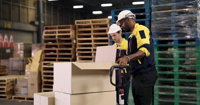 Multiracial male warehouse team lifting cardboard boxes onto pallet jack for cargo shipment while managing inventory in industrial storage facility with organized pallet stacks