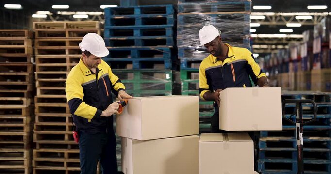 Multiracial warehouse workers collaborating on cardboard box packaging process during cargo preparation in storage area surrounded by wooden pallets and shrink-wrapped stock items