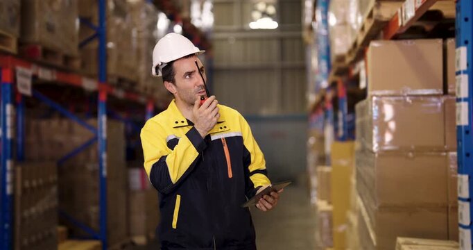 Caucasian male warehouse supervisor holds tablet and stands beside stacked cardboard boxes while controlling inventory data in organized stock facility with digital barcode system and safety gear