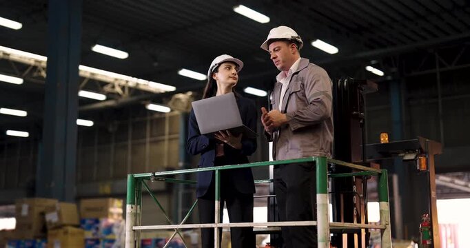 Warehouse supervisor and female manager discuss cargo data on laptop while standing on elevated forklift platform inside industrial distribution center with safety gear and pallet stacks in background
