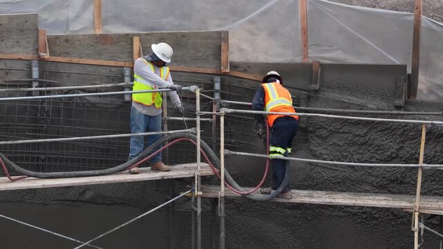 Construction workers on construction site scaffolding, wearing PPE performing shotcrete blasting or shooting concrete from a nozzle onto steel rebar.